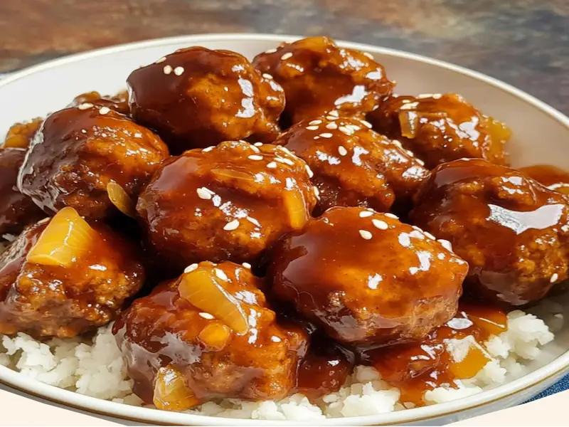 Bowl of sweet and sour meatballs topped with sesame seeds and green onions, served with rice.
